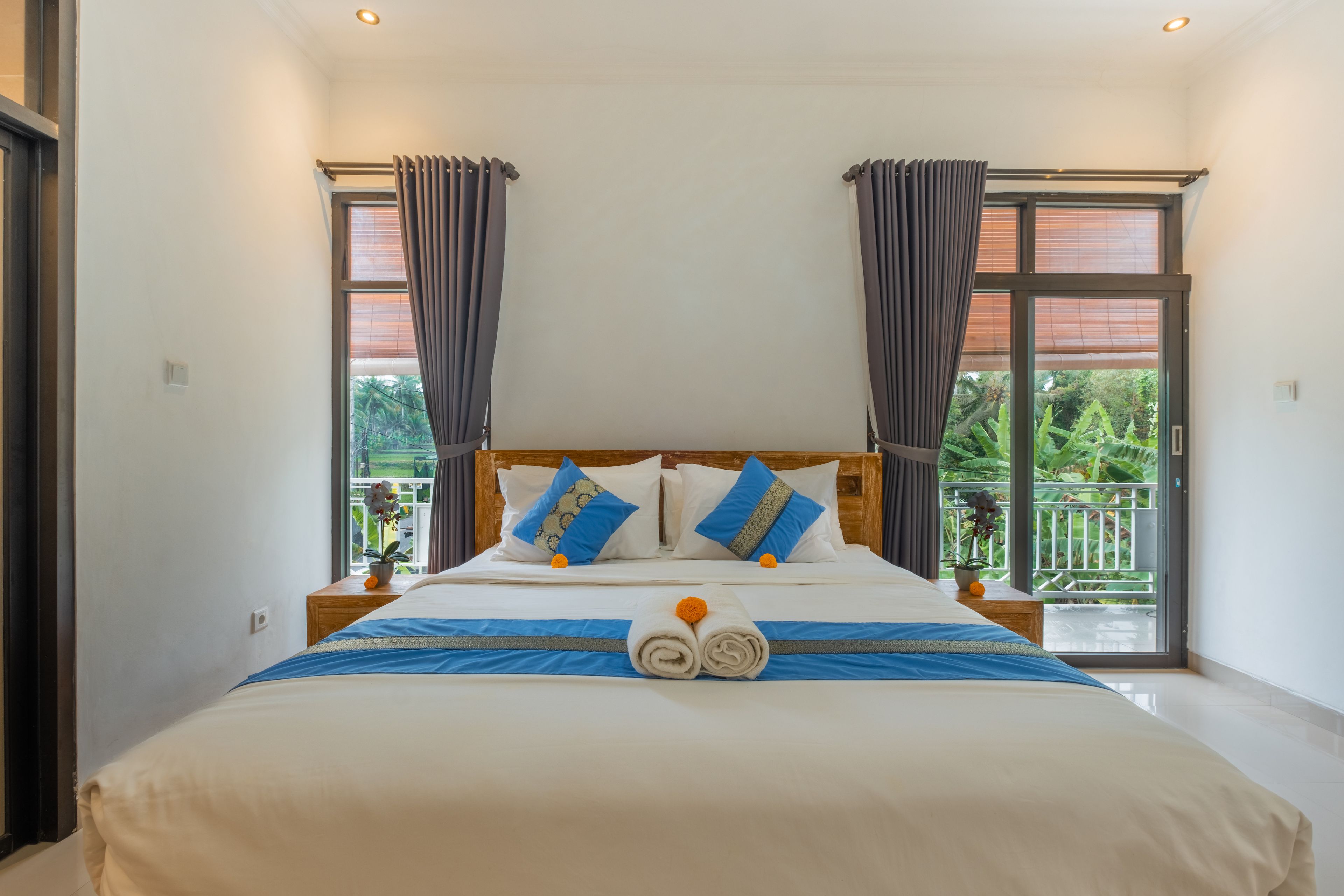 A hotel room with a large bed, two nightstands, a wooden headboard, and a balcony with a view of greenery.