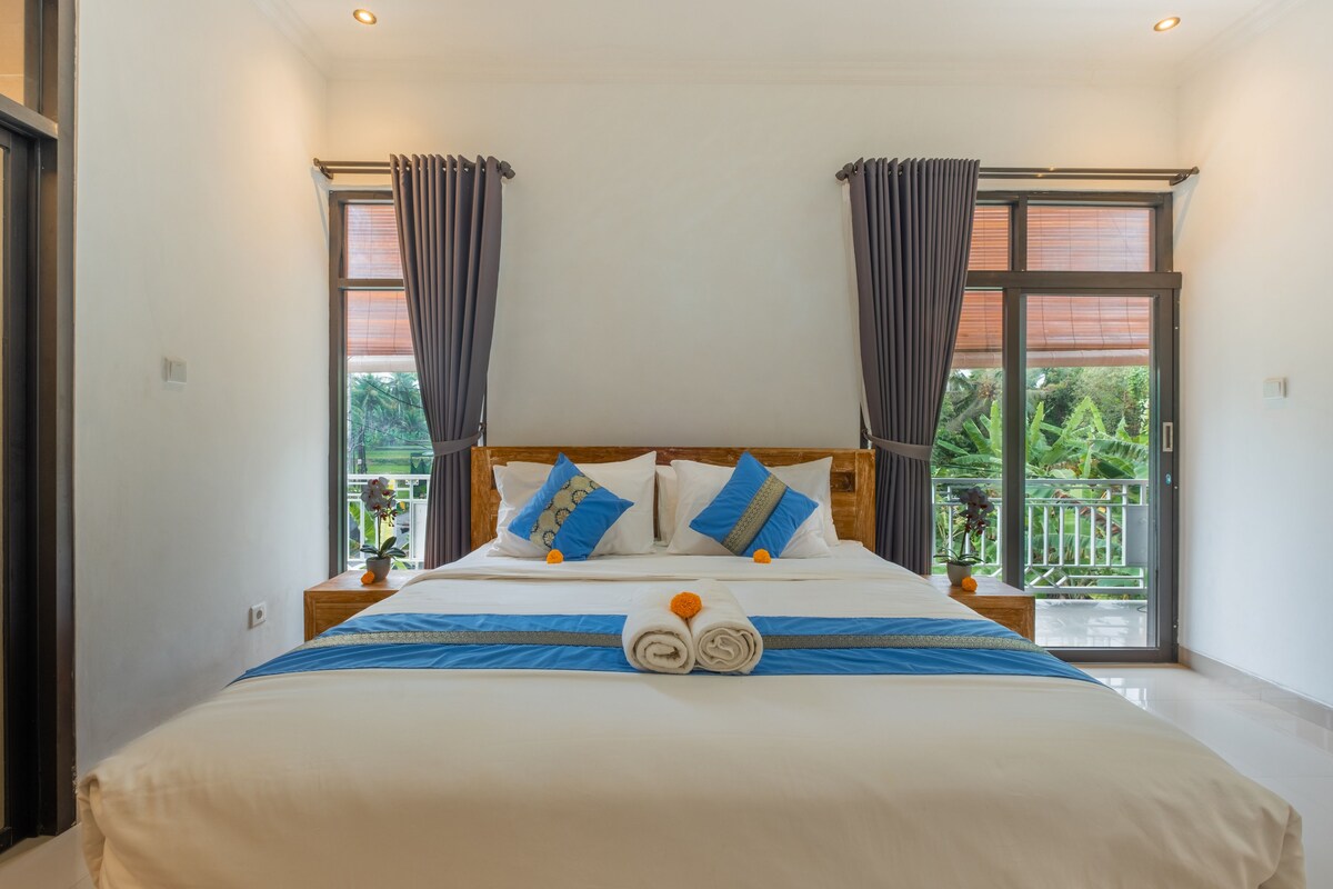 Habitación de hotel con una cama grande, dos mesitas de noche, cabecera de madera y un balcón con vistas a zonas verdes.