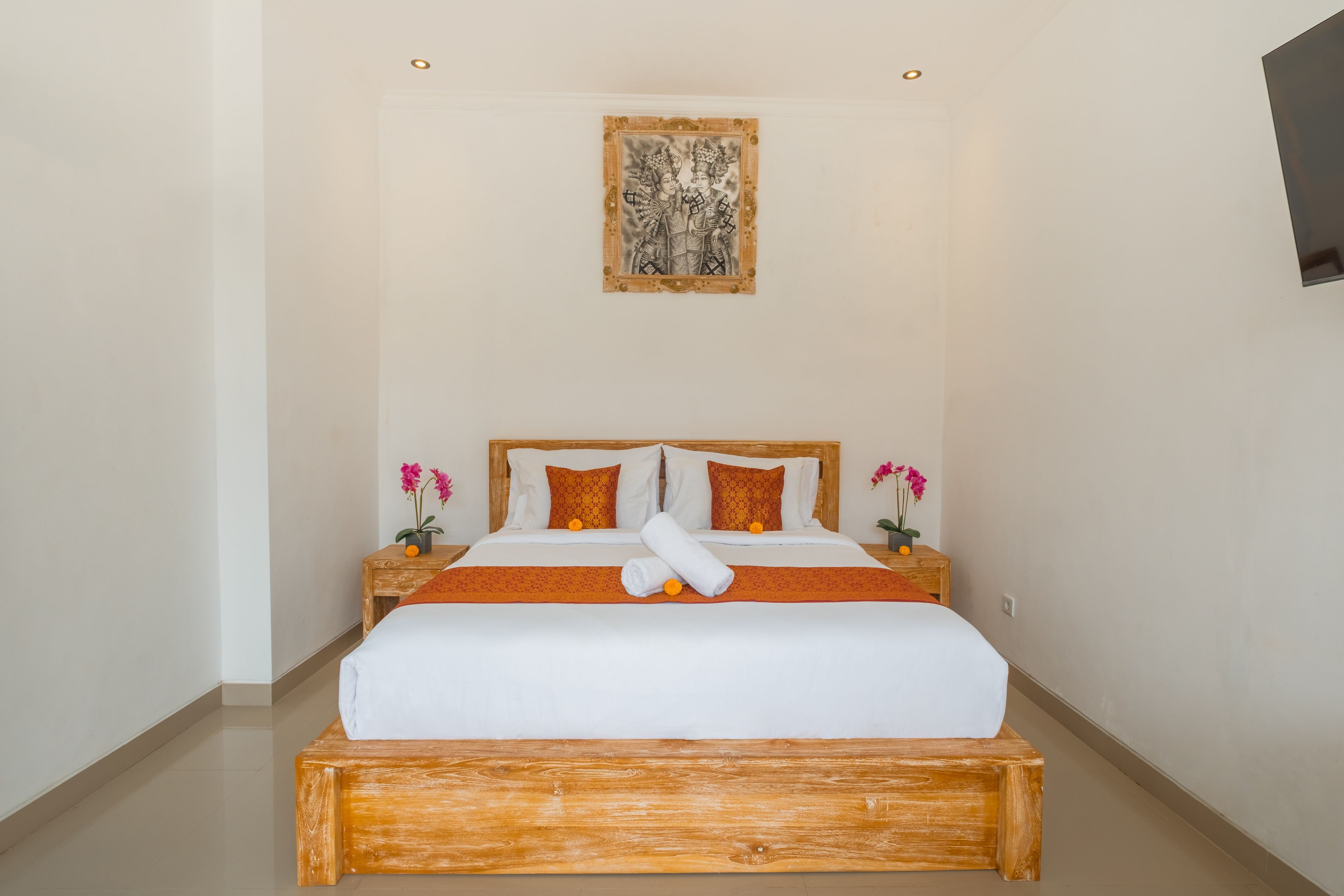 A hotel room with a bed, wooden headboard, two nightstands with flower vases, and a painting above the bed.