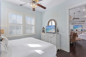 A bedroom with a large bed, a ceiling fan, a TV, and a view of a beach through a window. - Luxury Prominence Retreat | Bikes, Resort Pool & Walk to The Big Chill (Watersound)