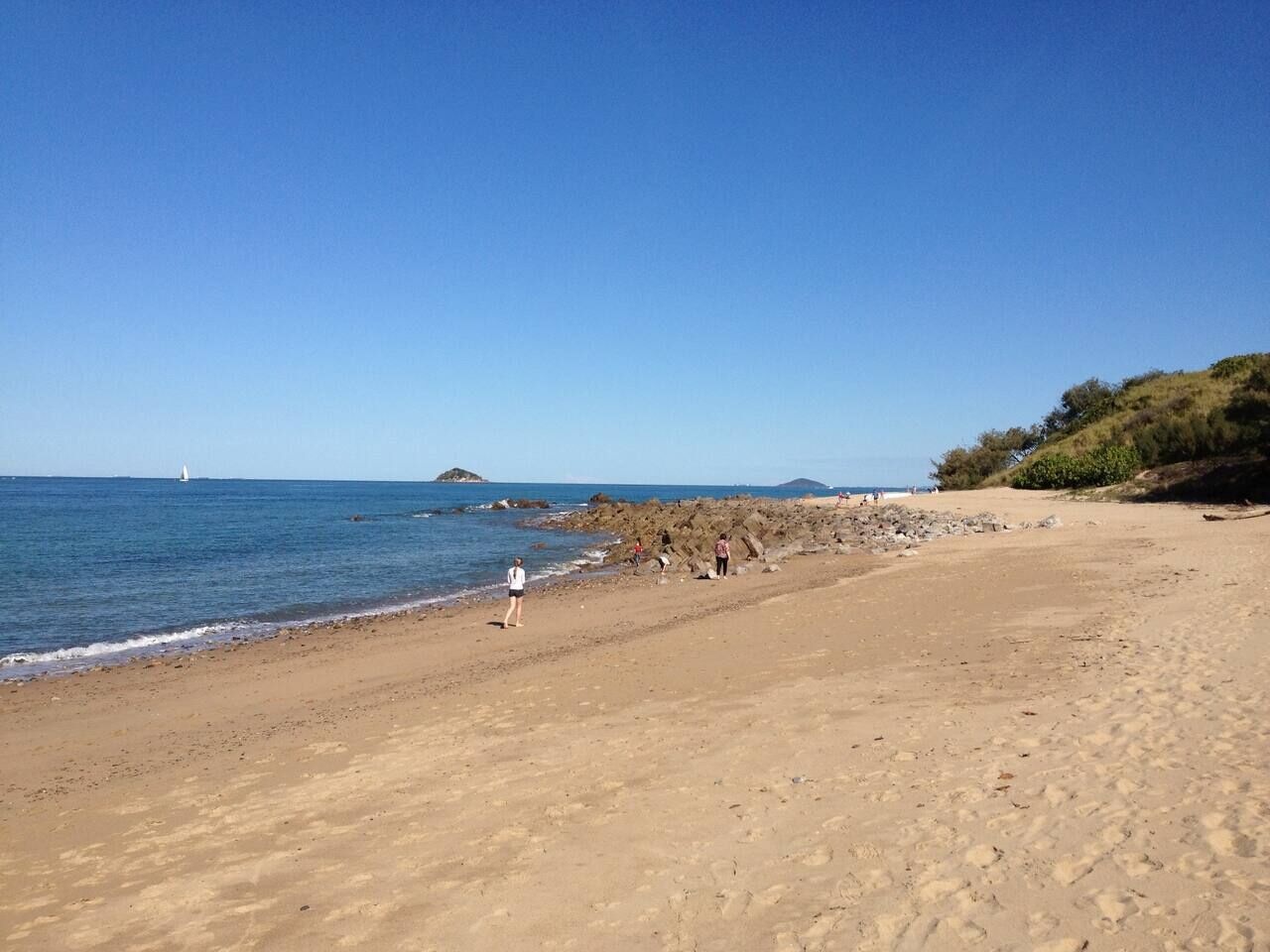 A sandy beach with a clear blue sky, a distant sailboat, and rocky outcrops.