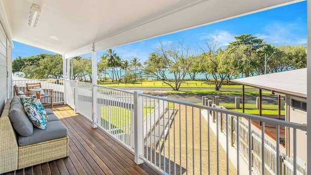 A covered outdoor area with a wooden deck, white railing, and a view of a park with trees and a body of water.