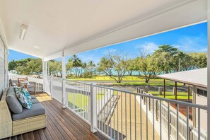A covered outdoor area with a wooden deck, white railing, and a view of a park with trees and a body of water.