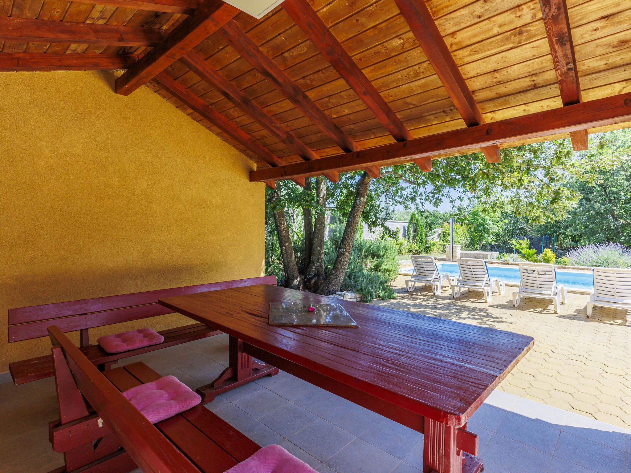 Un espace pique-nique abrité, avec une table et des bancs en bois, entouré de verdure et d’une piscine.