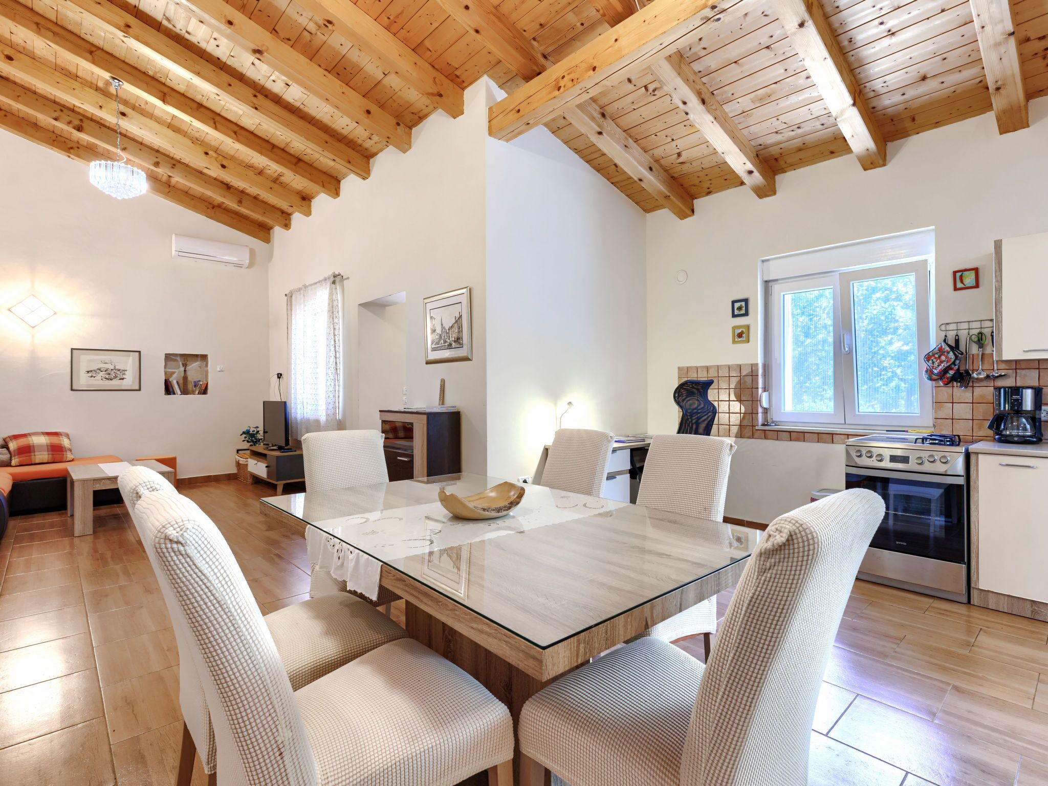 A cozy living area with a wooden ceiling, a dining table with chairs, a TV, and a kitchen with a stove and oven.