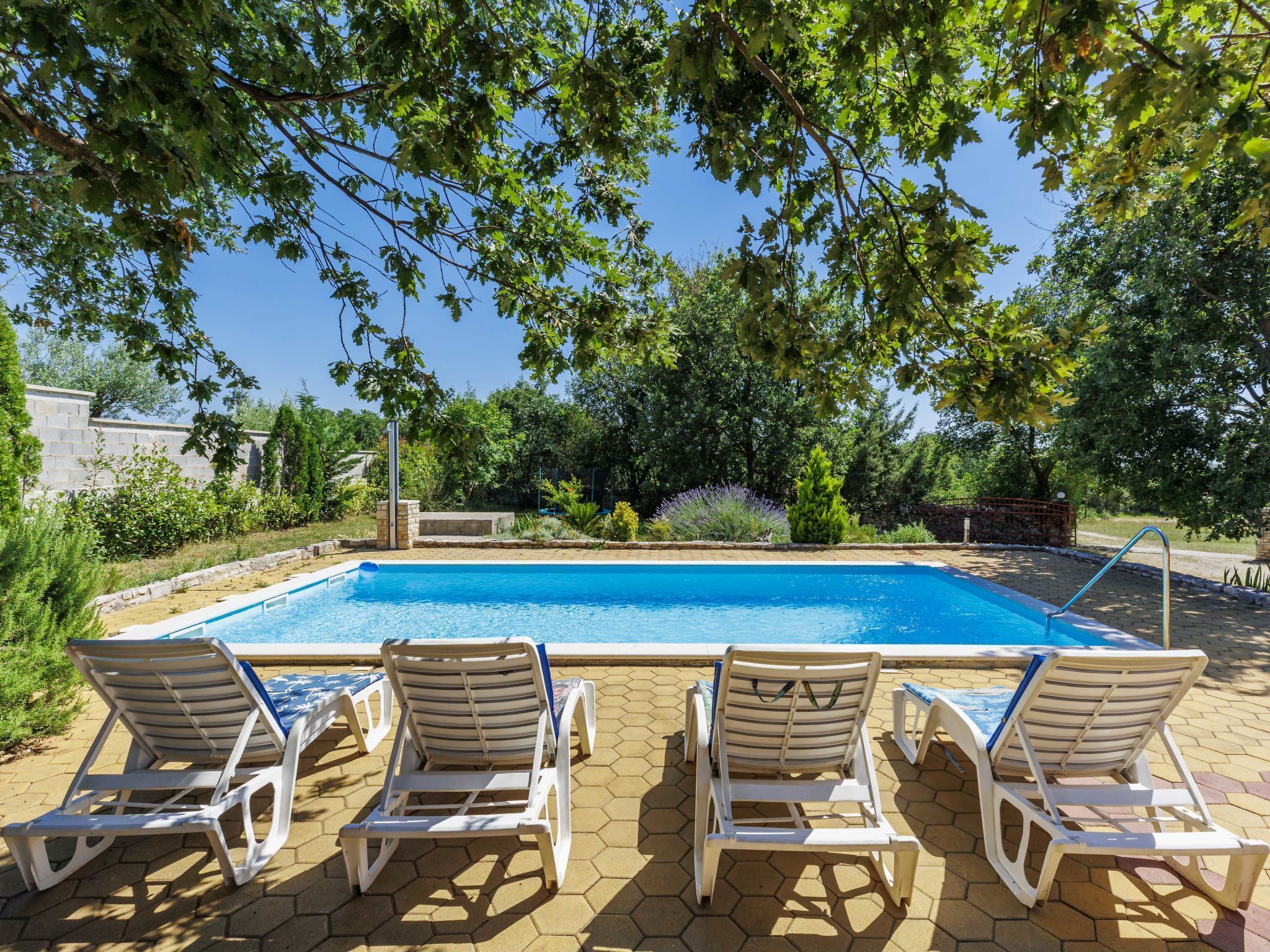 A rectangular swimming pool with clear blue water, surrounded by a tiled patio with hexagonal tiles. There are four white lounge chairs with blue cushions aligned along the pool's edge.