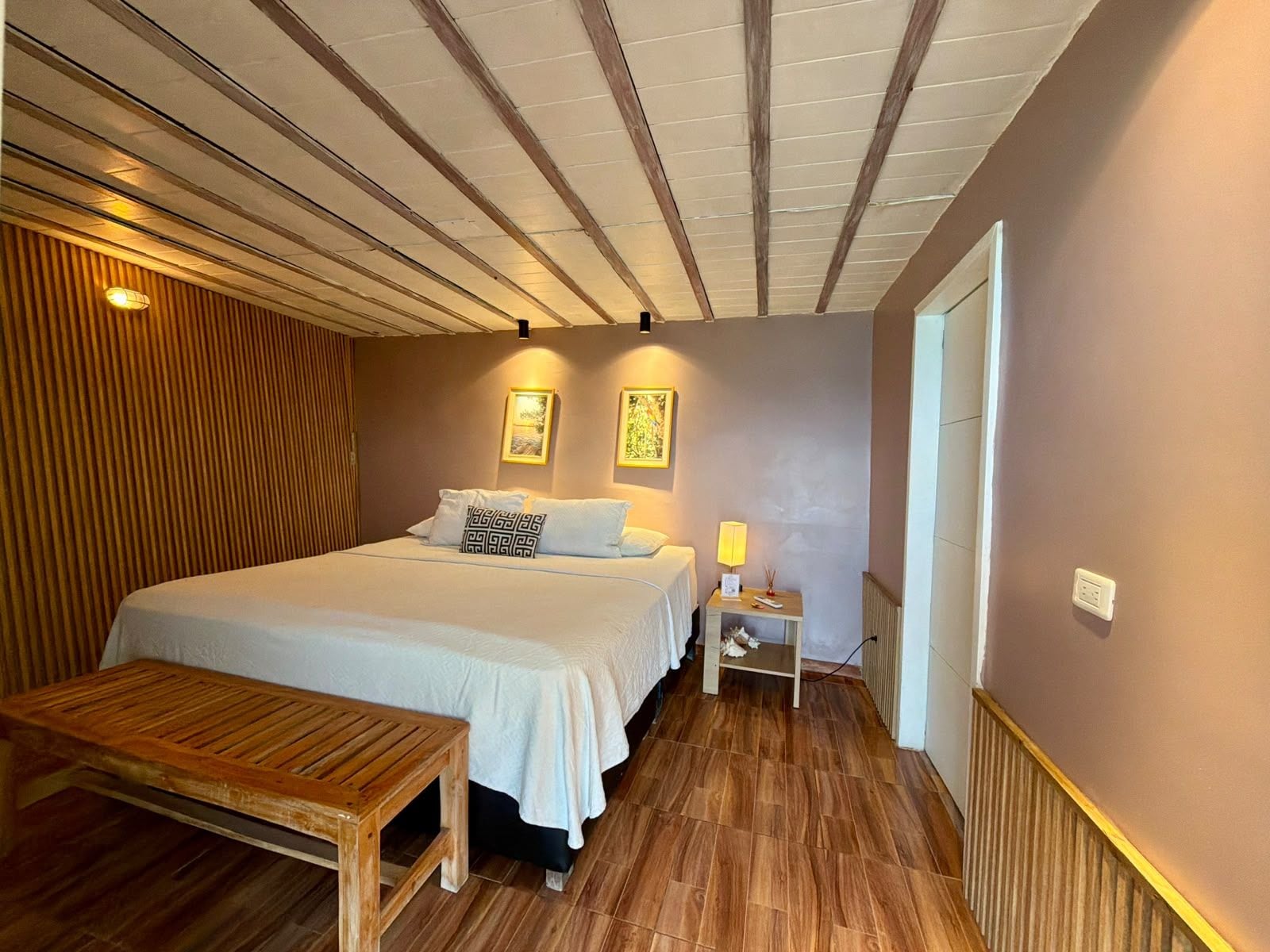 A bedroom with a wooden floor, a bed with white bedding, a bedside table with a lamp, and a bench.