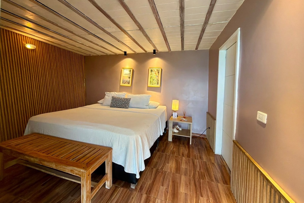 Quarto com piso de madeira, cama com roupa de cama branca, mesa de cabeceira com abajur e um banco.