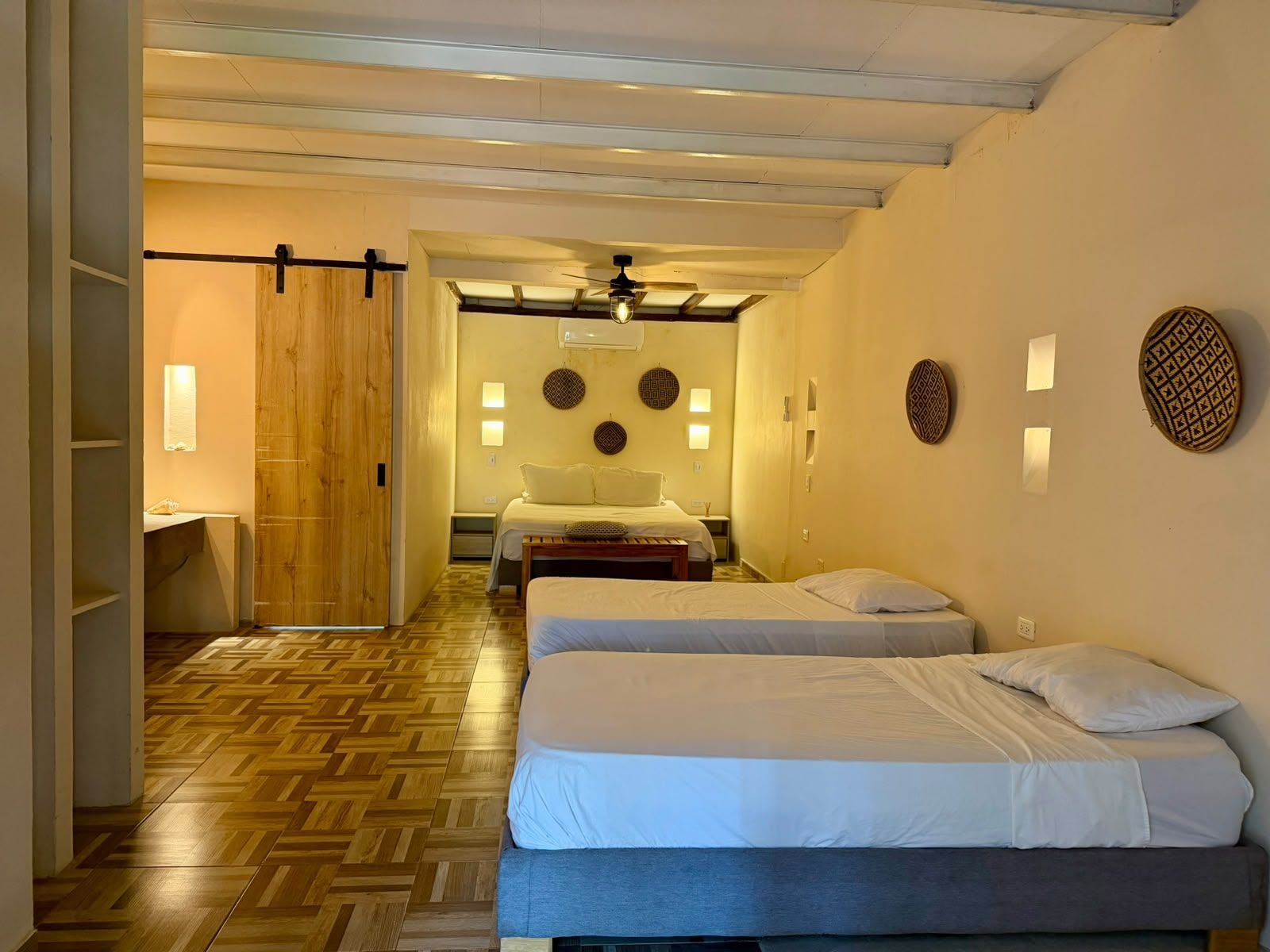 A hotel room with two beds, a wooden floor, a sliding door, and wall-mounted lights.