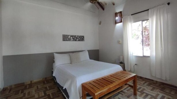 A bedroom with a bed, a wooden bench, a nightstand, a wall-mounted light, and a window with curtains. - Raices del rosario  (Rosario Islands)
