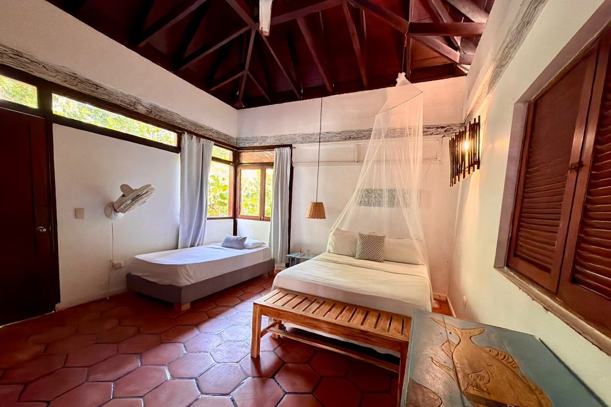 Quarto com cama, um banco, janela com cortinas e luminária na parede.