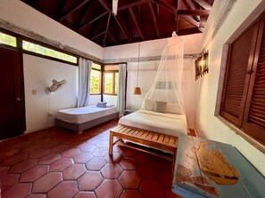 Una habitación con cama, una banca, una ventana con cortinas y una lámpara de pared.