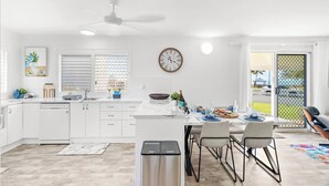 Une cuisine moderne avec des meubles blancs, un coin repas comprenant une table et des chaises, un ventilateur de plafond et une vue à travers une porte-fenêtre coulissante.