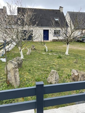 Ein Haus mit dunklem Dach und blauer Tür, umgeben von einer grünen Rasenfläche mit Bäumen und großen Steinen.