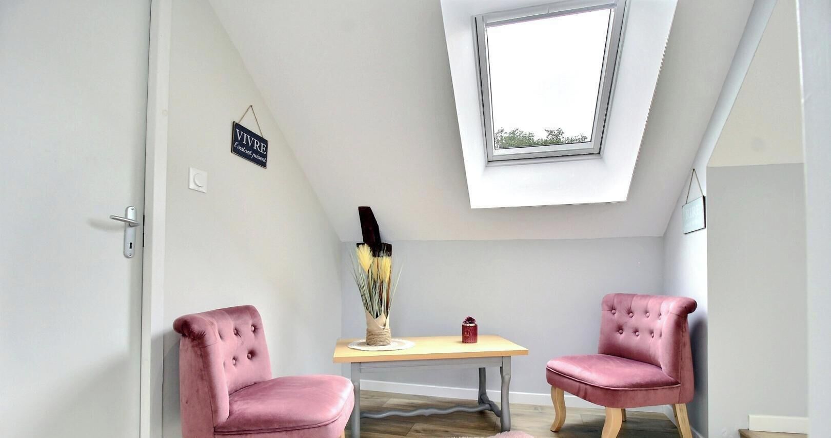 A room with a sloped ceiling, a skylight, two pink chairs, a small table with a vase and a candle, and a sign on the wall.