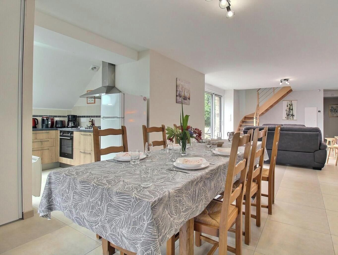 A dining area with a table set for four, wooden chairs, a kitchen in the background, and a staircase leading to another level.