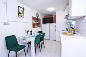 Una cuina compacta amb armaris blancs, una zona de menjador amb taula preparada per a dues persones, i un televisor muntat sobre l'armari de la nevera.