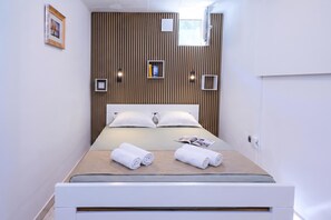 Moderni hotellihuone, jossa on puupanelilla verhoiltu seinä, valkoisilla lakanoilla varustettu sänky, kaksi rullalle taitettua pyyhettä ja lehti sängyllä.