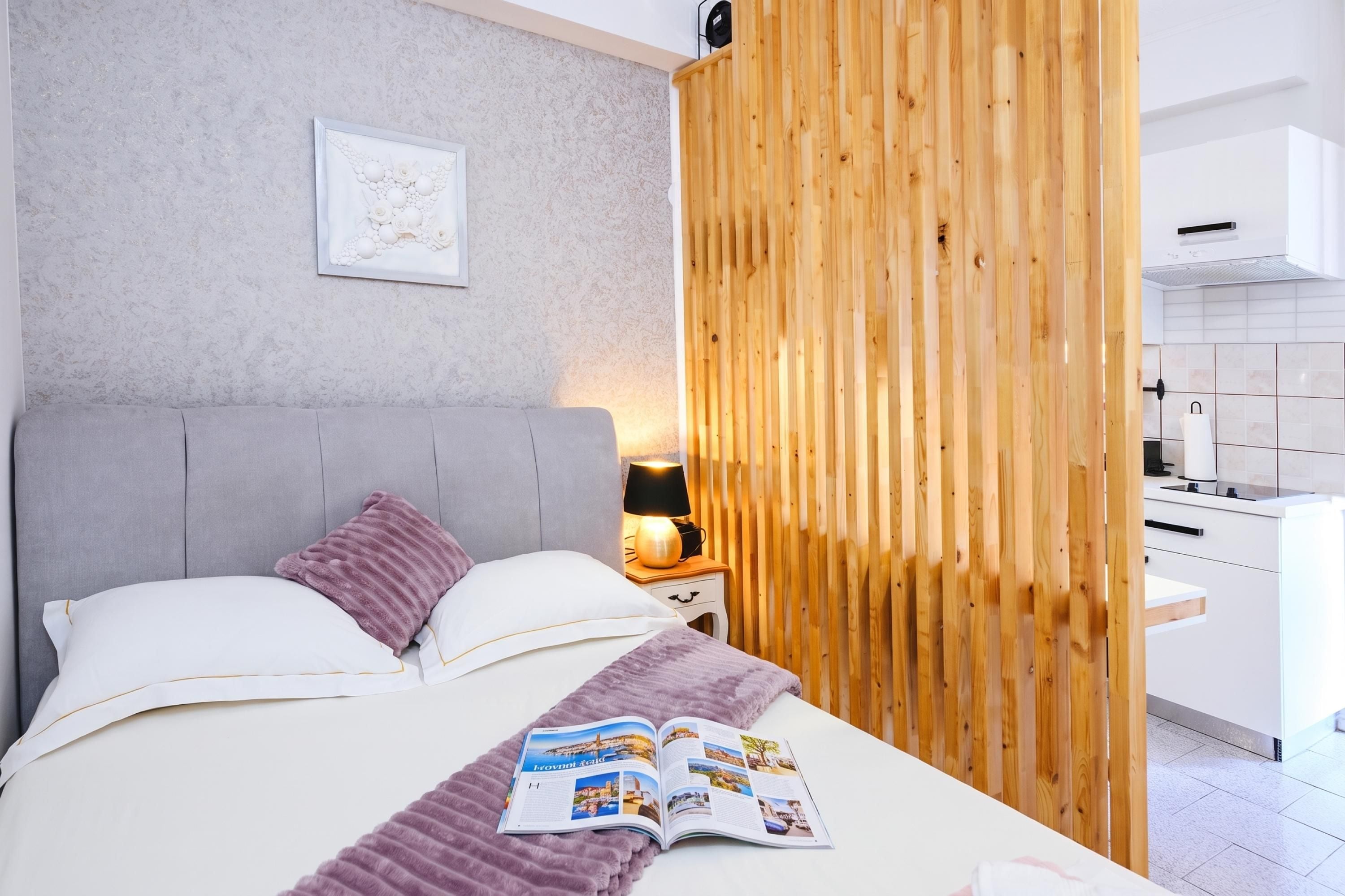 A modern bedroom with a bed, pillows, a bedside table with a lamp, a magazine, and a wooden partition.