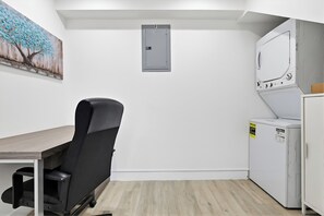 Een kamer met een bureau, een stoel, een wasmachine en een schilderij aan de muur.