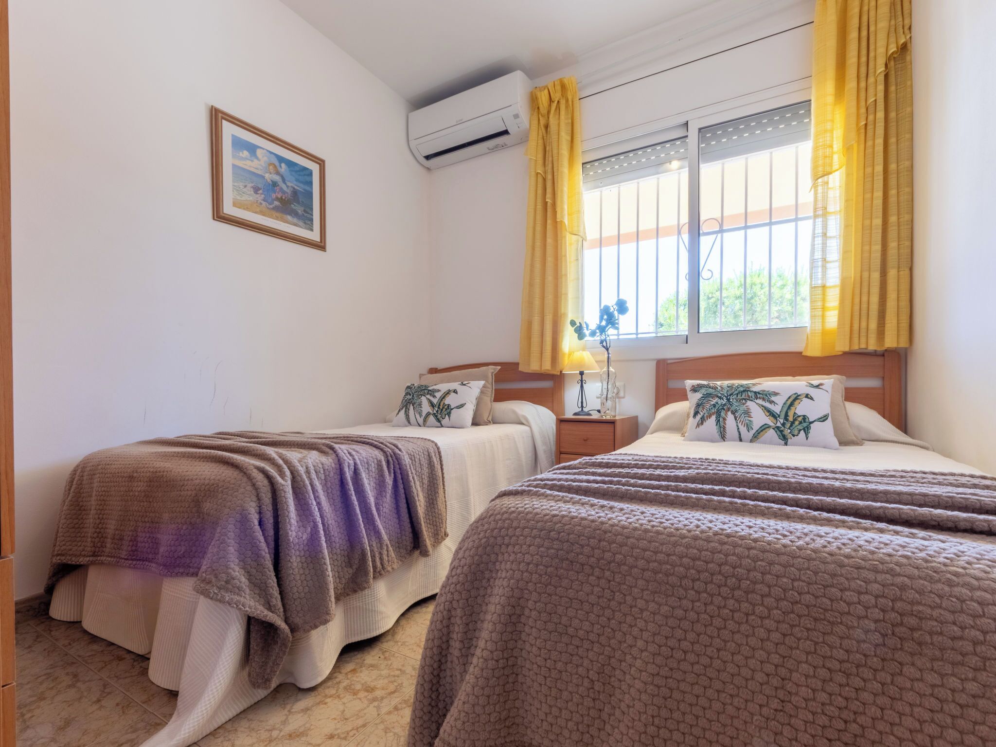 Une chambre d’hôtel avec deux lits, une fenêtre avec des rideaux jaunes, un climatiseur fixé au mur et un tableau encadré.