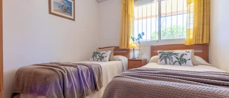 A hotel room with two beds, a window with yellow curtains, a wall-mounted air conditioner, and a framed picture.