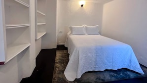Habitación estrecha con cama, paredes blancas y una lámpara en el techo.