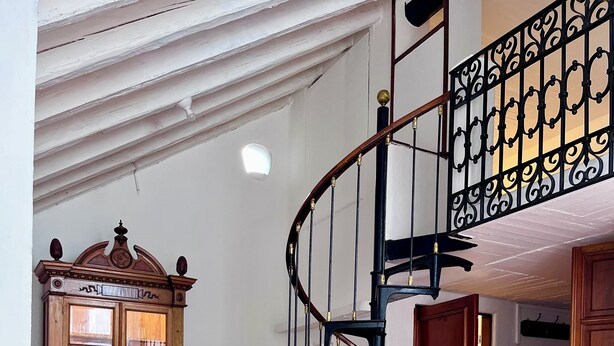 Una escalera en espiral con barandilla negra, una silla de madera con cojín y un aparador con puertas de cristal.