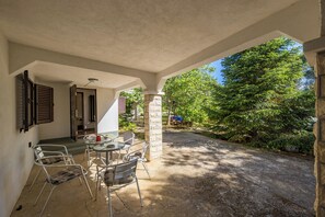 Un patio cubierto con una mesa y sillas, un pilar de piedra y vista a un camino bordeado de árboles.
