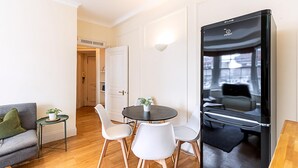 A modern room with a round table, white chairs, a grey sofa, a small side table with a plant, and a black refrigerator. - Modern Flat Near Hyde Park (London)