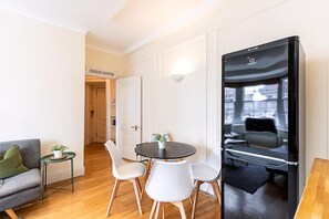 Ein modernes Zimmer mit einem runden Tisch, weißen Stühlen, einem grauen Sofa, einem kleinen Beistelltisch mit Pflanze und einem schwarzen Kühlschrank.
