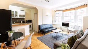A modern living room with a sofa, a dining table with chairs, a TV, and a kitchen area with cabinets and a microwave. - Modern Flat Near Hyde Park (London)