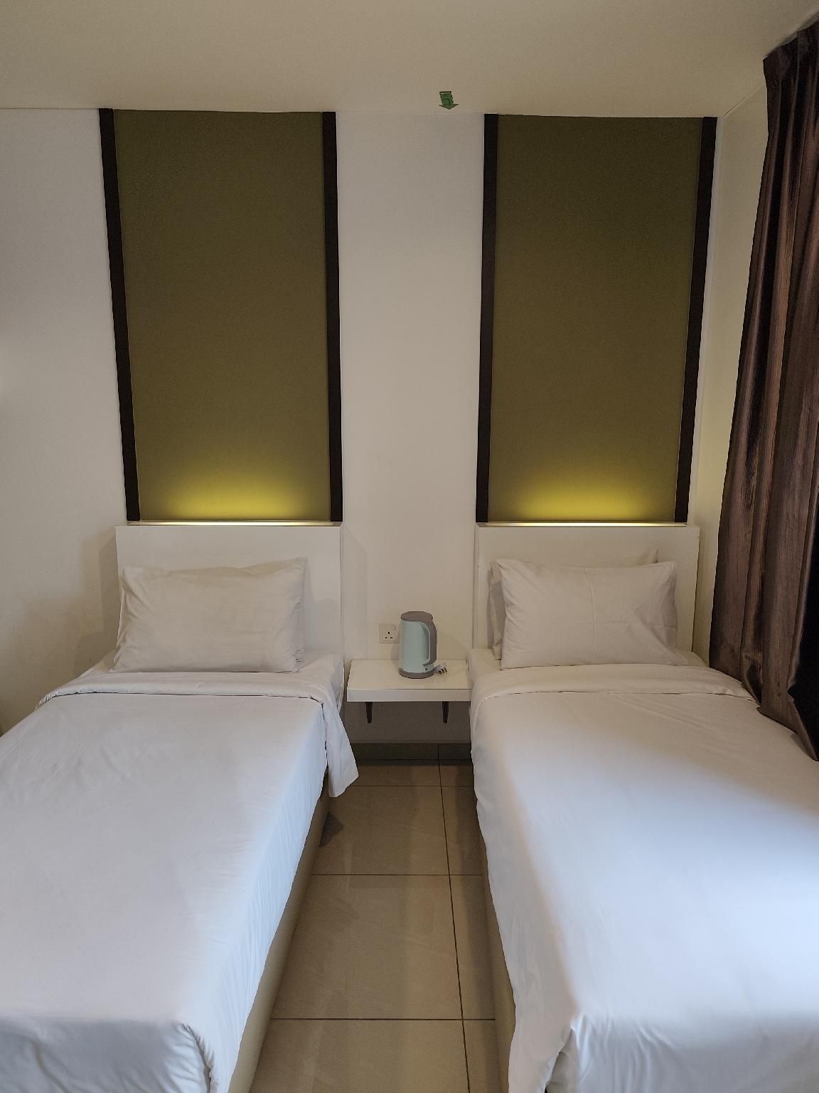 Habitación de hotel con dos camas individuales, una mesita de noche pequeña con hervidor y una pared blanca con detalle marrón.