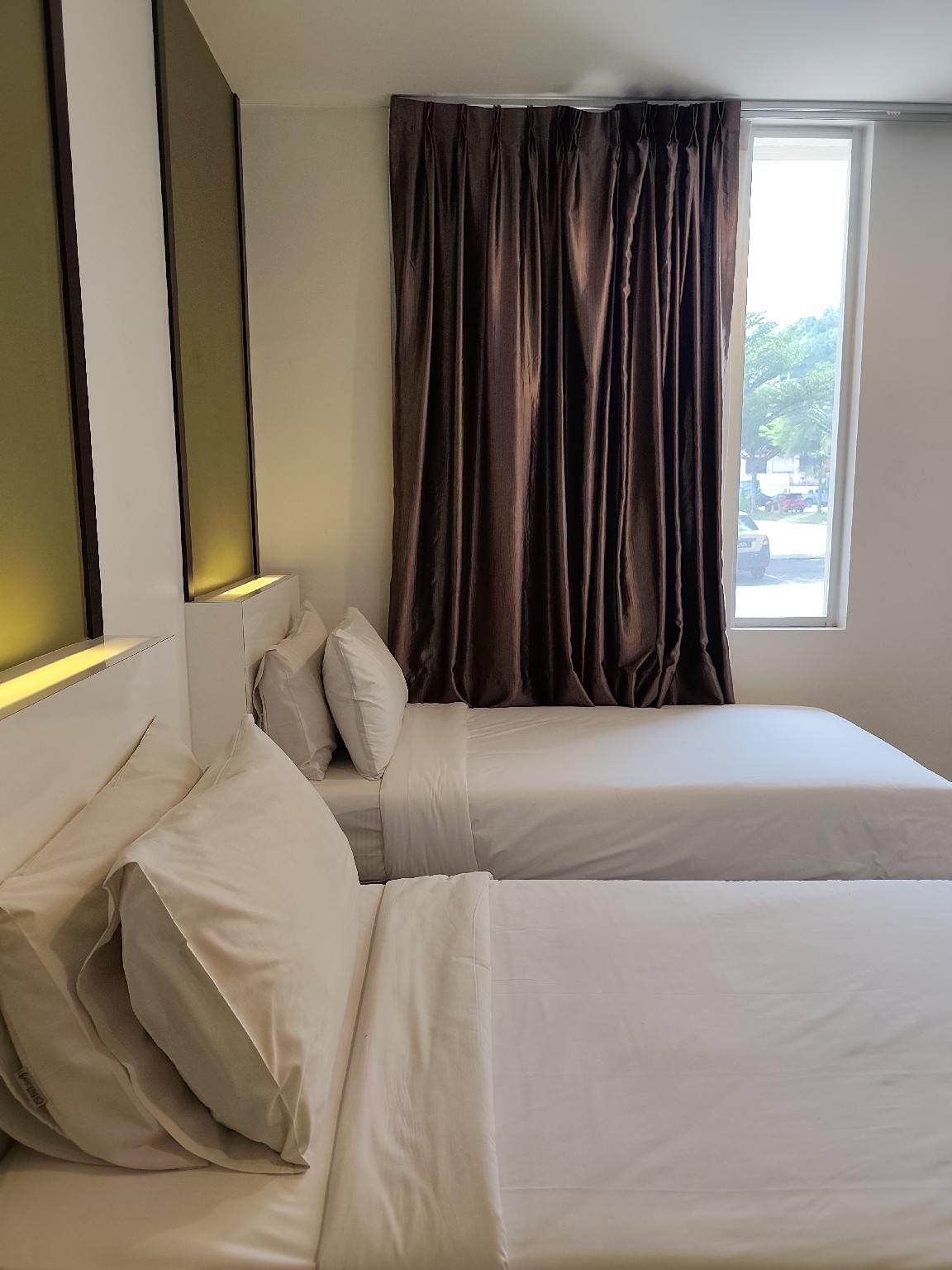A hotel room with a bed, pillows, a window with curtains, and a wall-mounted light fixture.