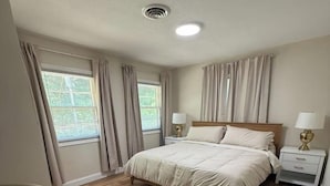 A bedroom with a wooden floor, a bed with white bedding, two bedside tables with lamps, and curtains on the windows. - The Reservoir House East (Andrews)