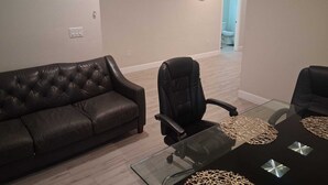A living room with a leather sofa, a black office chair, a glass coffee table with woven placemats, and a wall clock. - Serene 3-bedroom use in vibrant Orlando Close To Airport, Universal , Disney.... (Orlando)
