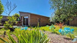 Een houten huis met een deur en ramen, omringd door een tuin met verschillende planten en een boom.