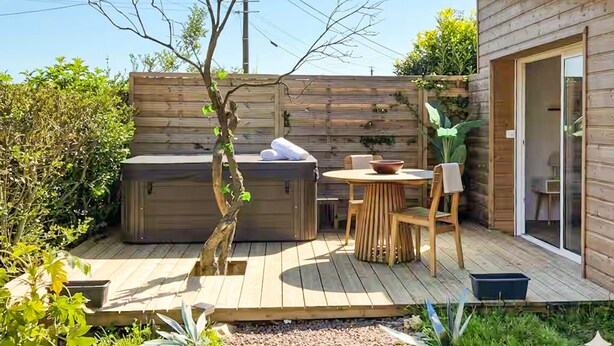 Een houten terras met een jacuzzi, een tafel met stoelen en een klein gebouw met een schuifdeur van glas.