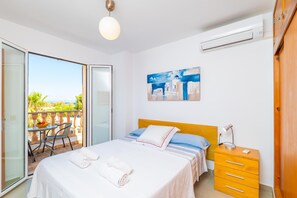 Una habitación de hotel con cama, mesita de noche, una silla, un balcón con mesa y sillas, y un cuadro en la pared.