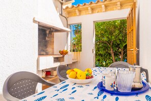 Un espacio de comedor al aire libre con una mesa preparada para dos, con mantel blanco y diseños de peces azules, un cuenco de fruta y una jarra de agua.