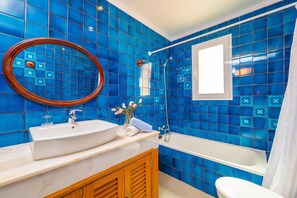 Un baño con azulejos azules, un lavamanos blanco, un armario de madera, una bañera blanca y una ventana con cortina blanca.