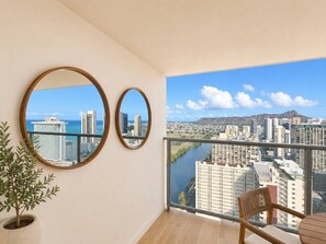 Ein Balkon mit Blick auf eine Stadtlandschaft, zwei runde Spiegel an der Wand, ein Blumentopf und ein Korbstuhl.