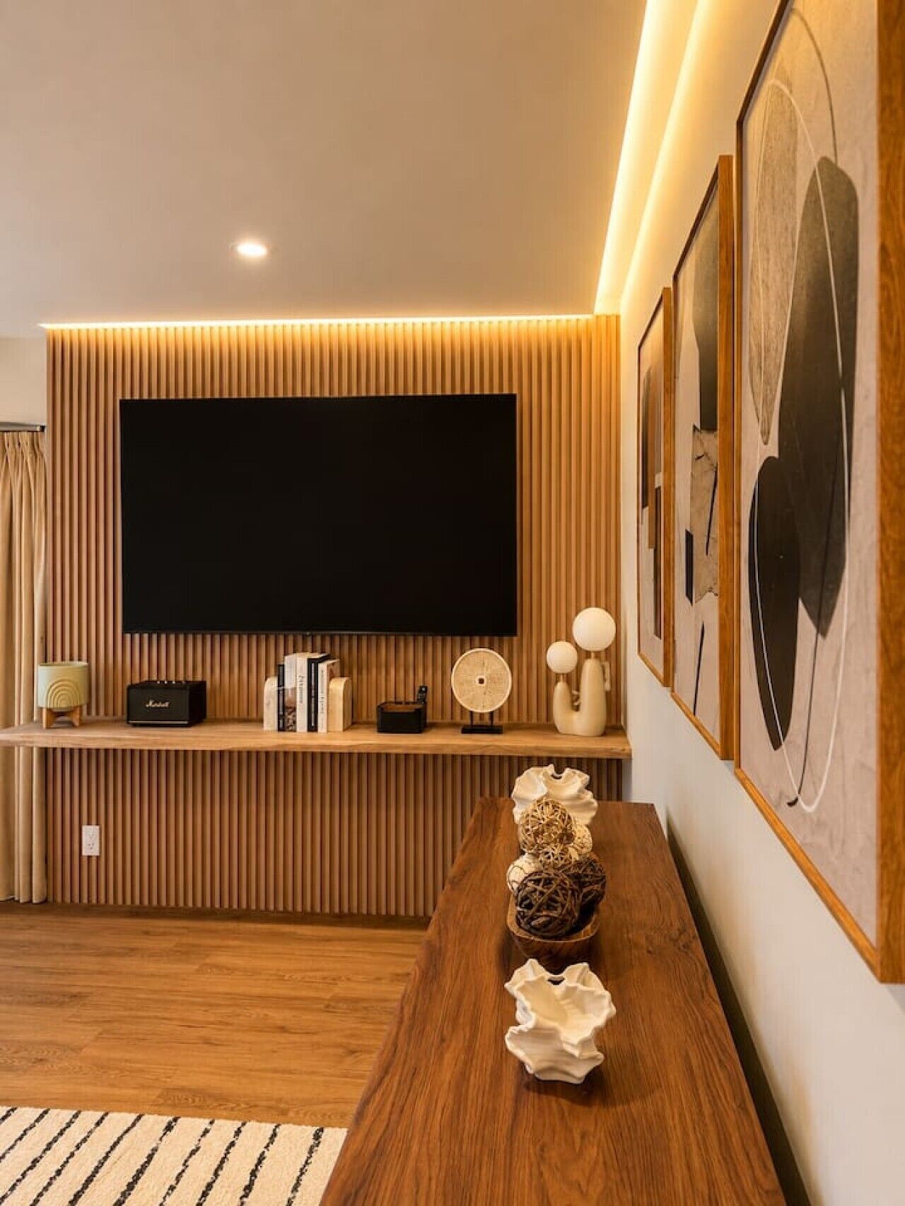 A modern living room with a large flat-screen TV mounted on a wooden wall, a wooden console table with decorative items, and framed artwork on the wall.