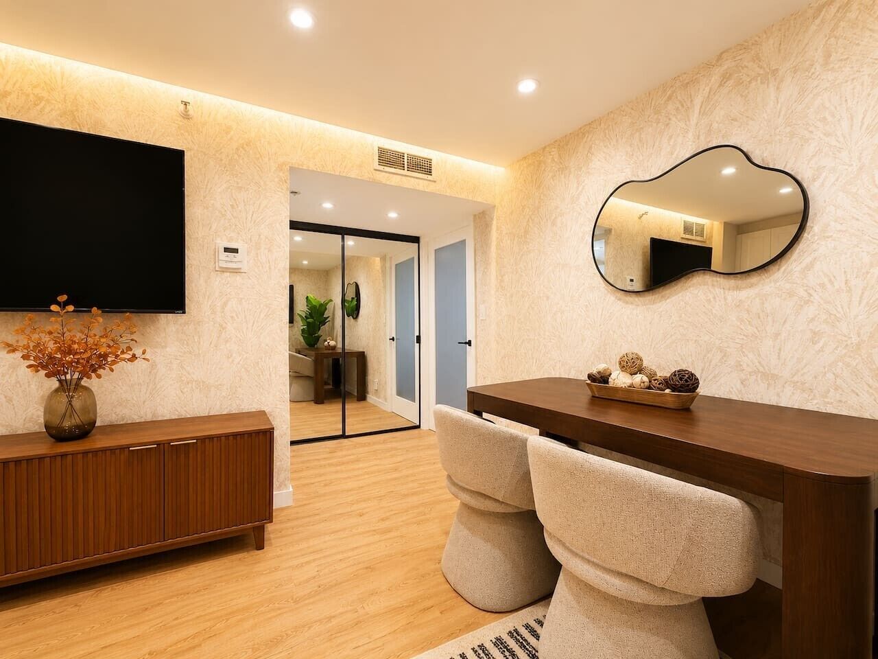 A modern hotel room with a wooden floor, a flat-screen TV mounted on the wall, a large mirror, and a wooden console table with a vase of dried flowers.