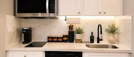 Una cocina con armarios blancos, microondas, cafetera, lavaplatos y fregadero.