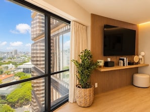 Ein modernes Hotelzimmer mit großem Fenster, einem Flachbildfernseher, einem Holz-TV-Möbel, einer Topfpflanze und Blick auf die Stadtlandschaft.