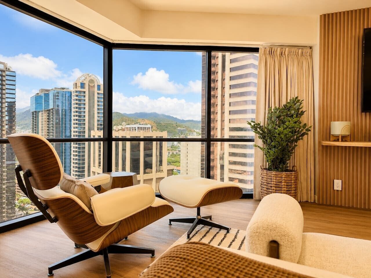 A modern hotel room with a large balcony, a TV, a potted plant, and a view of high-rise buildings.