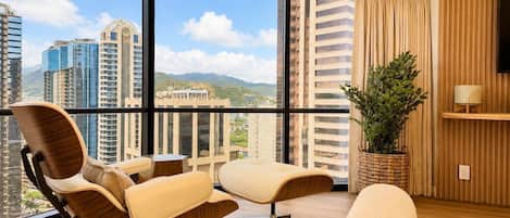 A modern hotel room with a large balcony, a TV, a potted plant, and a view of high-rise buildings.