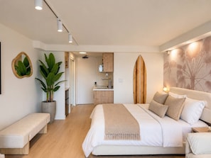 A modern bedroom with a large bed, bedside tables, a bench, a potted plant, and a kitchen area in the background. - Luxe Island Retreat – Modern Coastal Design (Honolulu)