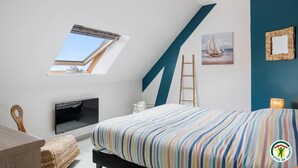 Een slaapkamer met schuine balken, een bed met een gestreept dekbed, een televisie, een raam met uitzicht, een ladder en een ingelijste foto aan de muur.